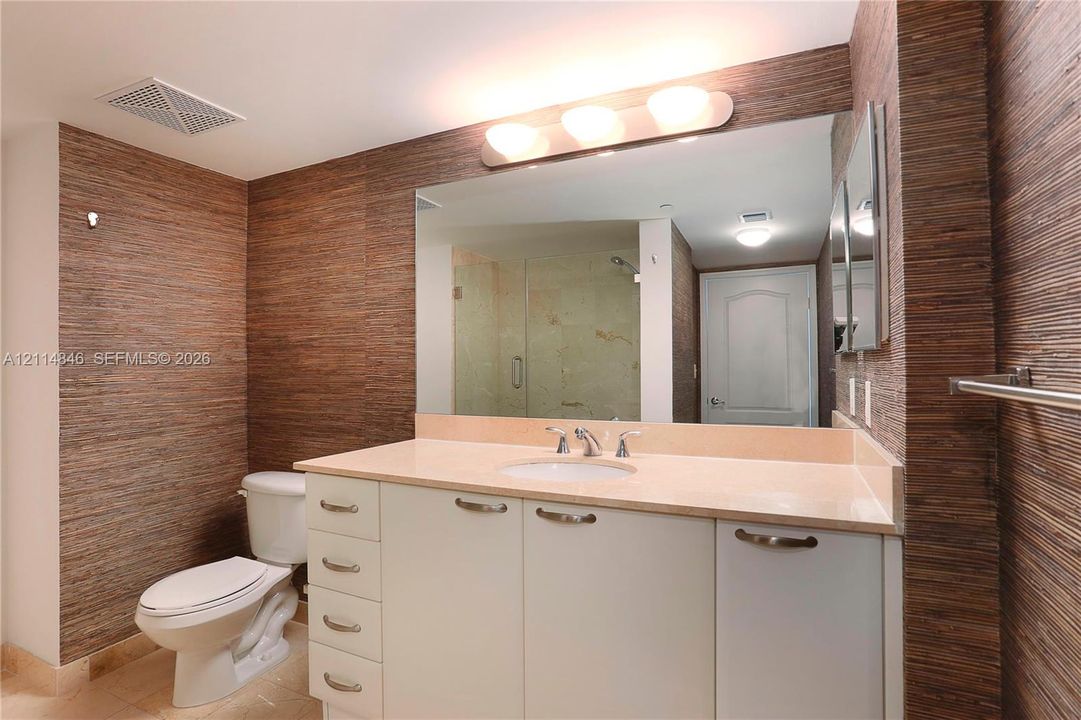 Bathroom featuring textured wall covering, a neutral-toned countertop with an integrated basin, white cabinetry with metallic hardware, and a large wall-mounted mirror