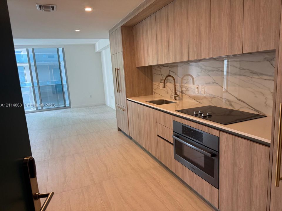 Kitchen featuring flat-panel wood-finish cabinetry, integrated appliances, light-toned stone-patterned backsplash, and ceramic tile flooring
