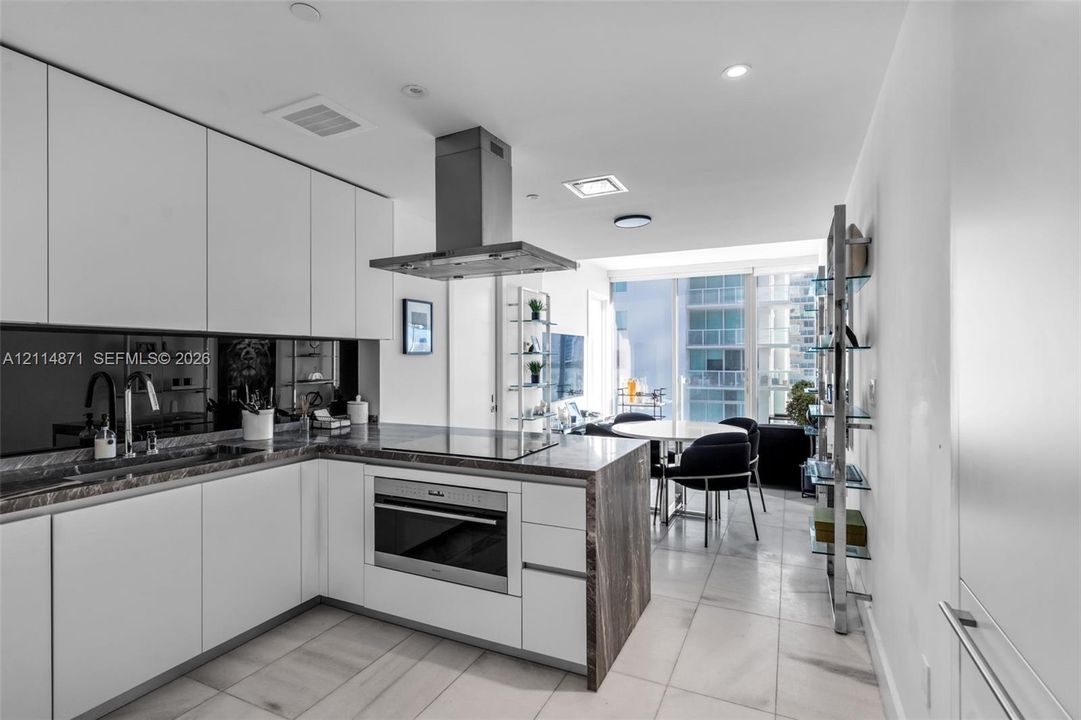 Contemporary kitchen featuring flat-panel white cabinetry, stone-surface countertops, a mirrored backsplash, built-in stainless steel oven, and an integrated stainless steel range hood