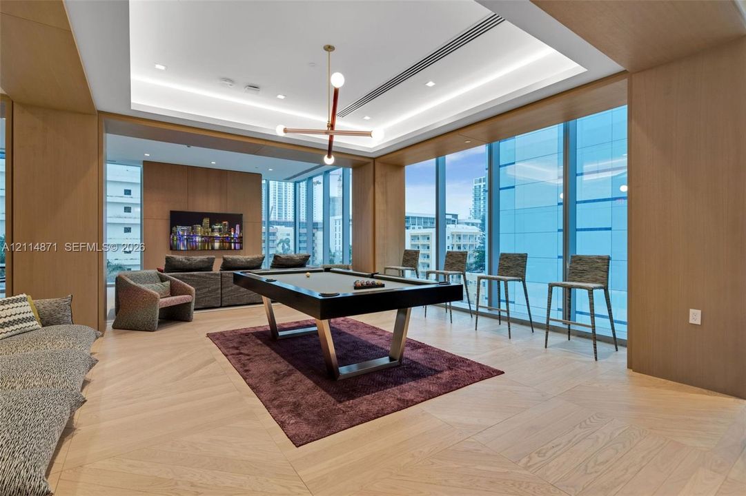 Floor-to-ceiling windows, wood-finish wall paneling, herringbone-patterned wood flooring, recessed tray ceiling lighting, and modern geometric chandelier