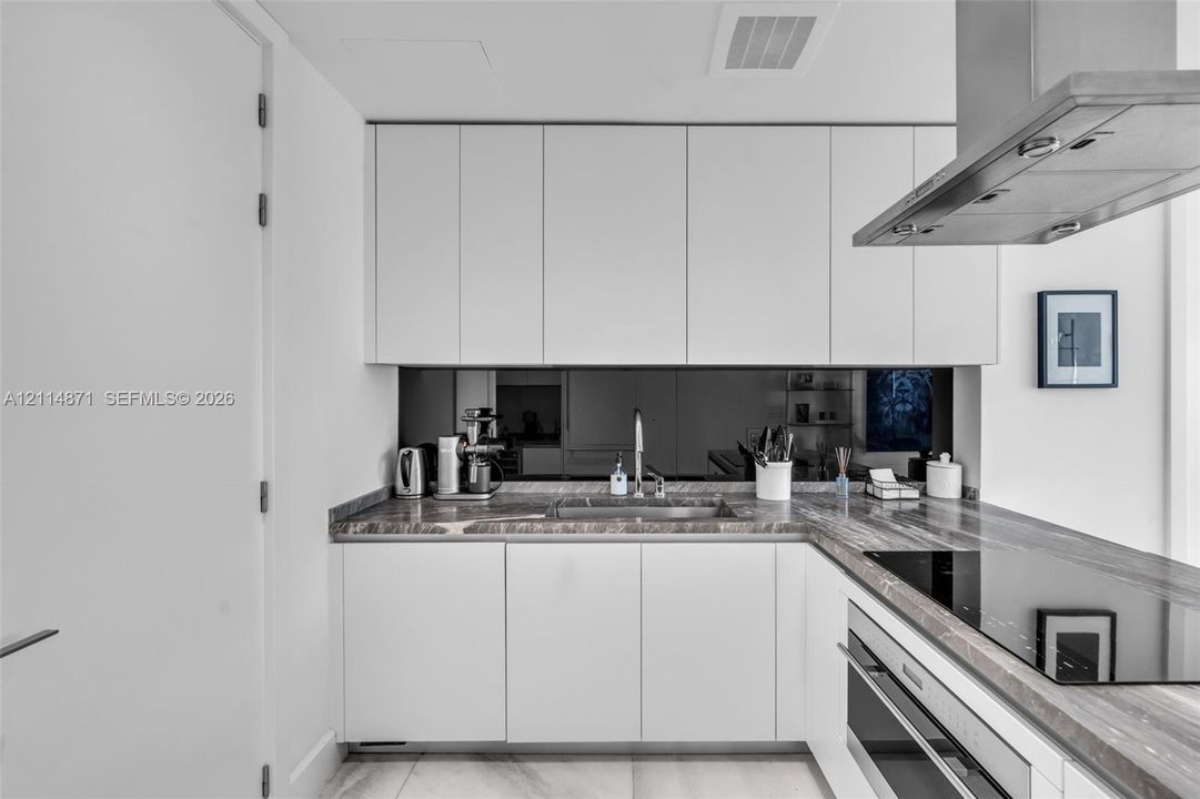 Modern kitchen featuring white flat-panel cabinetry, stone-finish countertops, mirrored backsplash, stainless steel range hood, and integrated cooktop