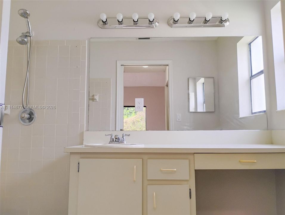 Bathroom vanity with a solid-surface countertop, drop-in sink, dual-fixture lighting, large wall-mounted mirror, and tile-surround shower with a handheld showerhead