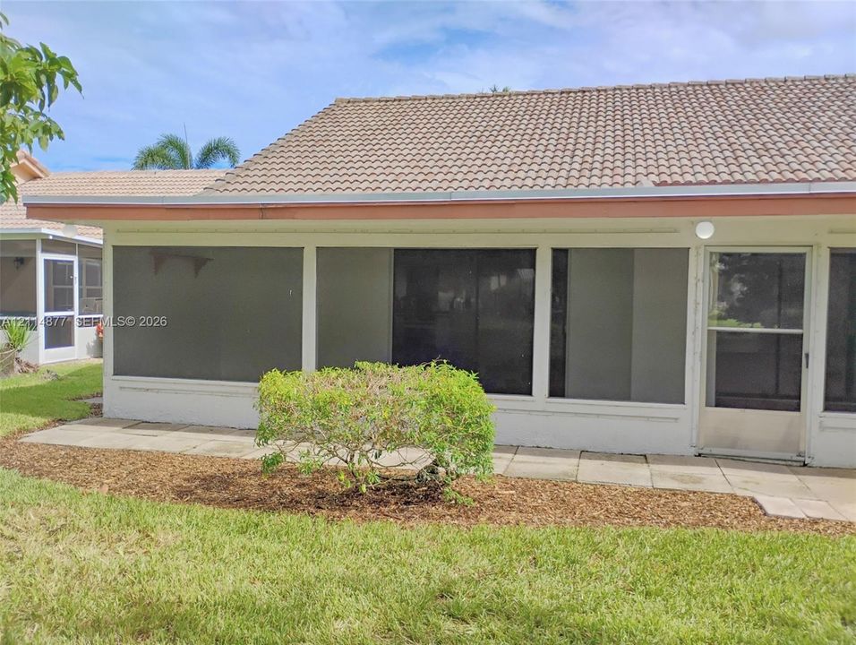Screened-in enclosure featuring a tiled roof, white exterior walls, and a dedicated screen door, set against a manicured lawn with mulched landscaping