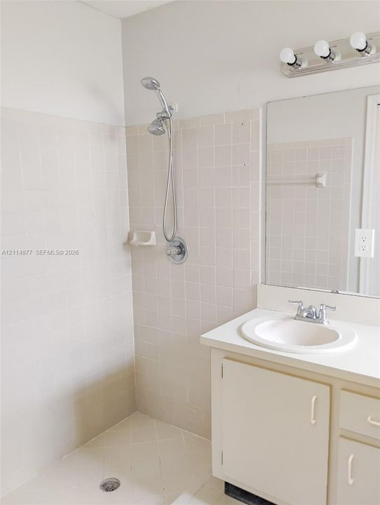 Bathroom featuring a tiled walk-in shower with a handheld showerhead, a vanity with an integrated basin, a wall-mounted mirror, and a three-bulb vanity light fixture