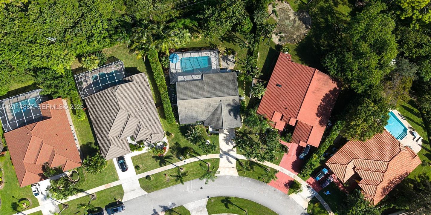 Aerial perspective of residential properties featuring concrete tile roofing, paved driveways, and screened-in swimming pools surrounded by mature trees