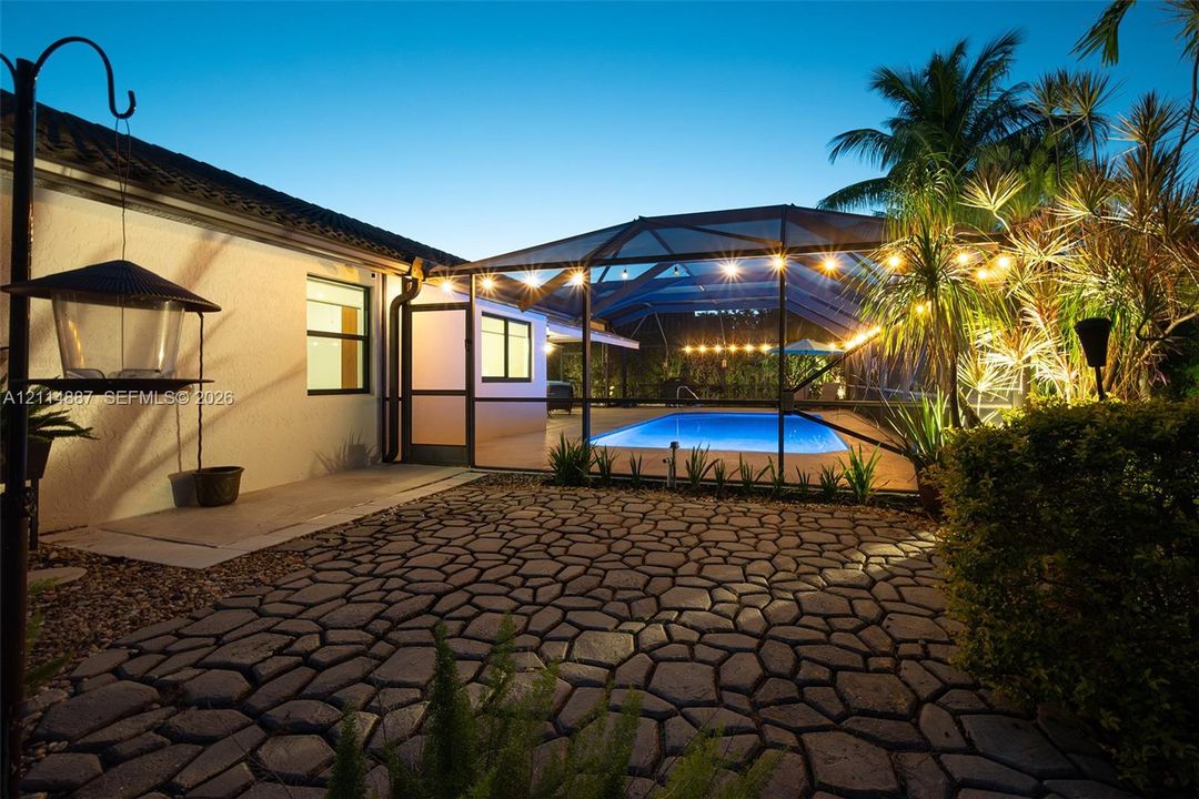 Screened-in outdoor pool area with stone-patterned paver patio, stucco exterior wall, and lush perimeter landscaping