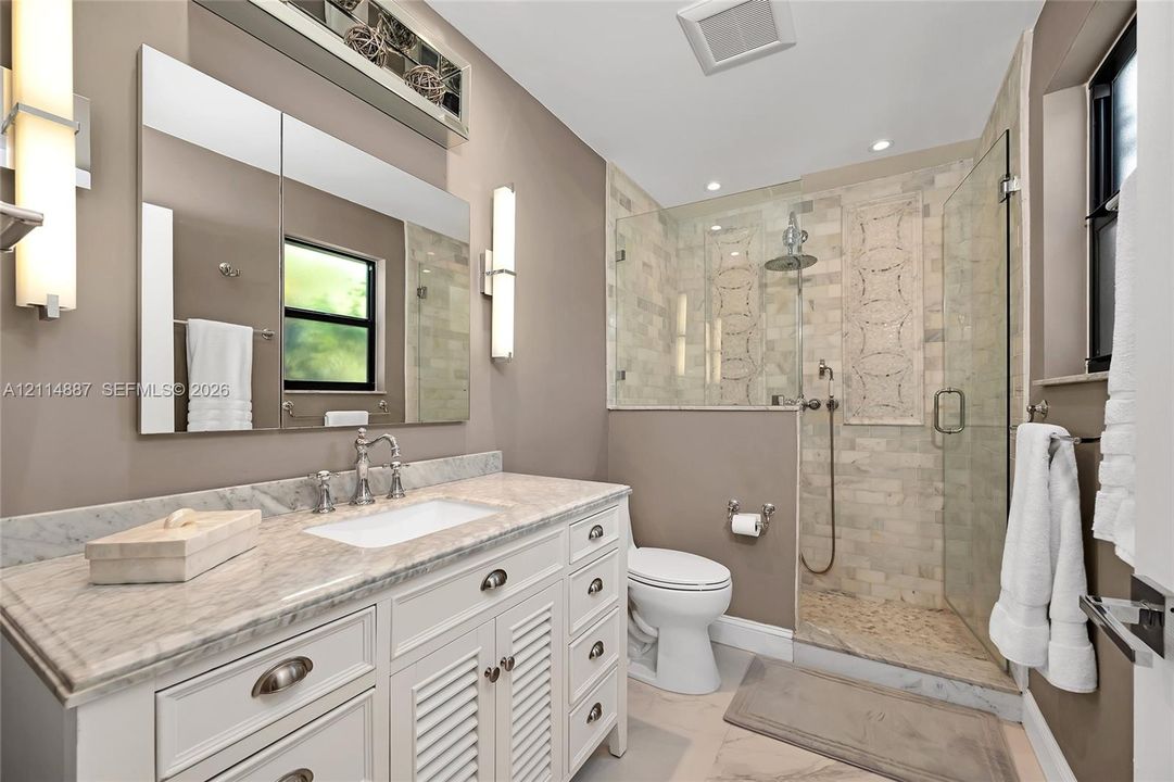 White vanity with stone countertop and undermount sink, walk-in shower with stone tile surround and decorative inlay, frameless glass enclosure, dual vertical wall sconces, and rectangular wall mirror