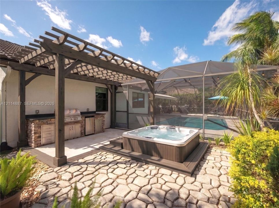 Outdoor kitchen featuring a stone-clad base, built-in stainless steel grill, and custom cabinetry beneath a dark timber pergola, complemented by an integrated spa and a screened-in swimming pool on a stone paver patio