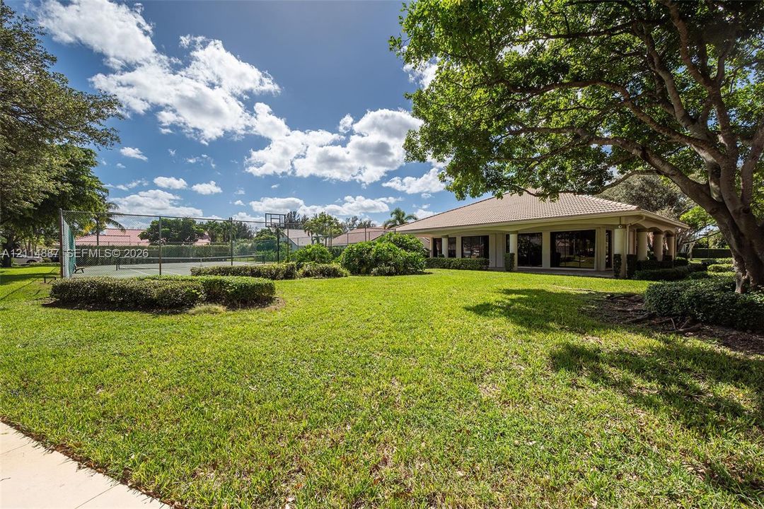 Expansive rear grounds featuring a fenced tennis court, manicured lawn, mature shade trees, and a covered patio