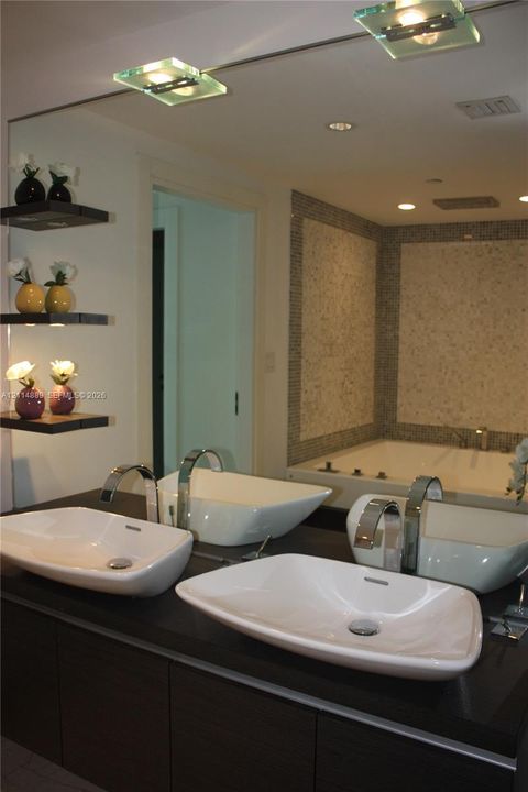 Dark wood-finish cabinetry topped with a dual-vessel sink vanity, featuring high-arc chrome faucets, wall-mounted display shelving, and an integrated jetted soaking tub with mosaic tile surrounds