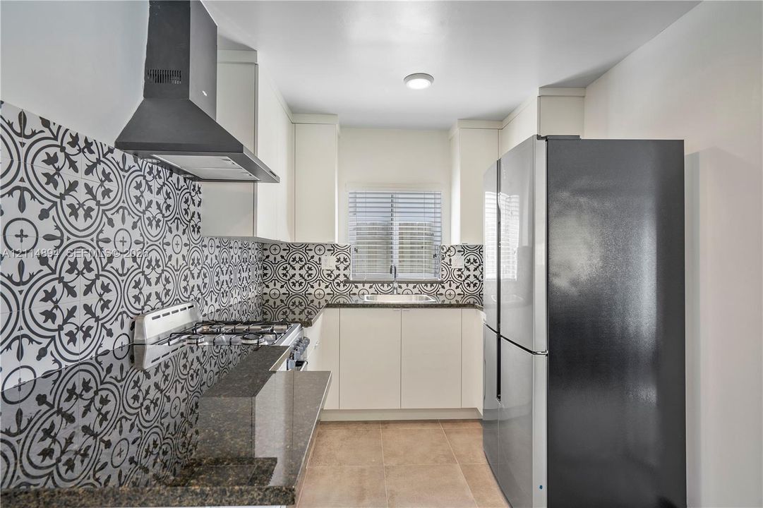 Kitchen featuring patterned tile backsplash, dark stone-finish countertops, matte black range hood, sleek white cabinetry, and stainless steel refrigerator
