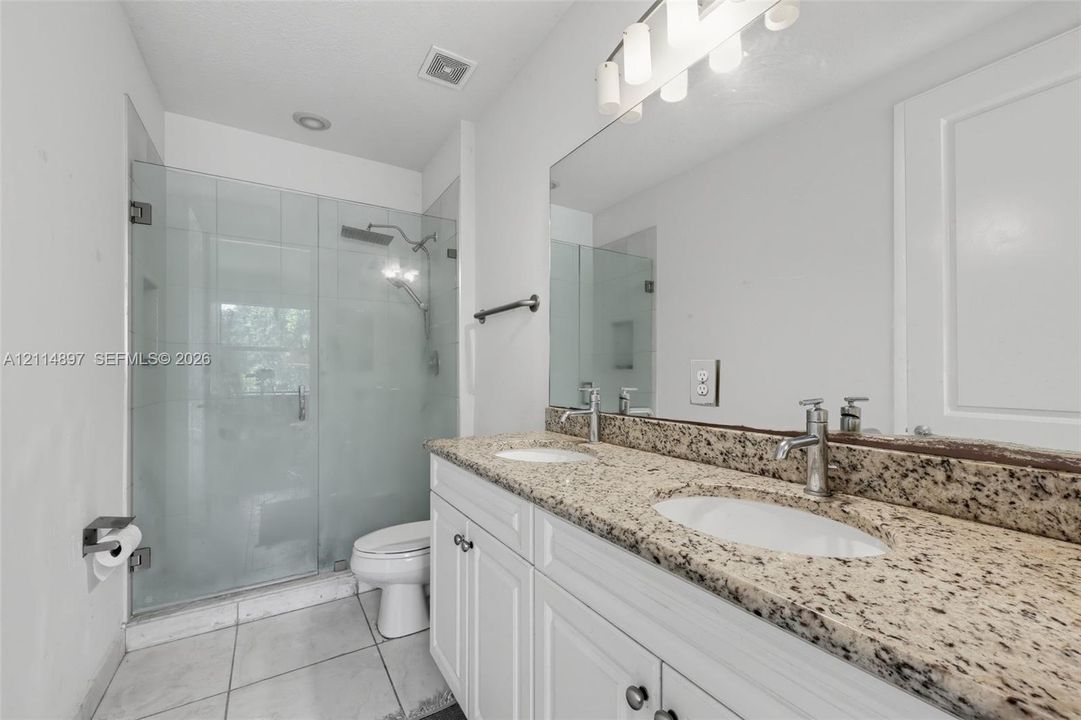 Dual-sink vanity with stone countertop and undermount basins, glass-enclosed shower, and tile flooring