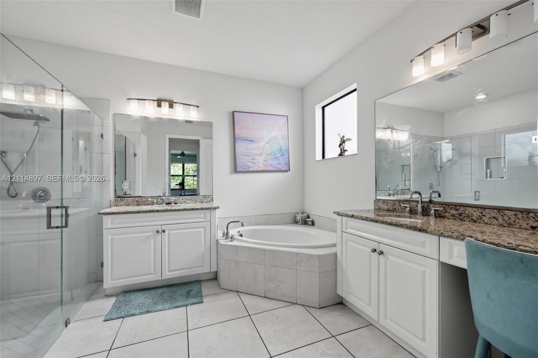 Dual white vanities with stone countertops, a glass-enclosed walk-in shower, a tiled corner soaking tub, and wall-mounted vanity lighting