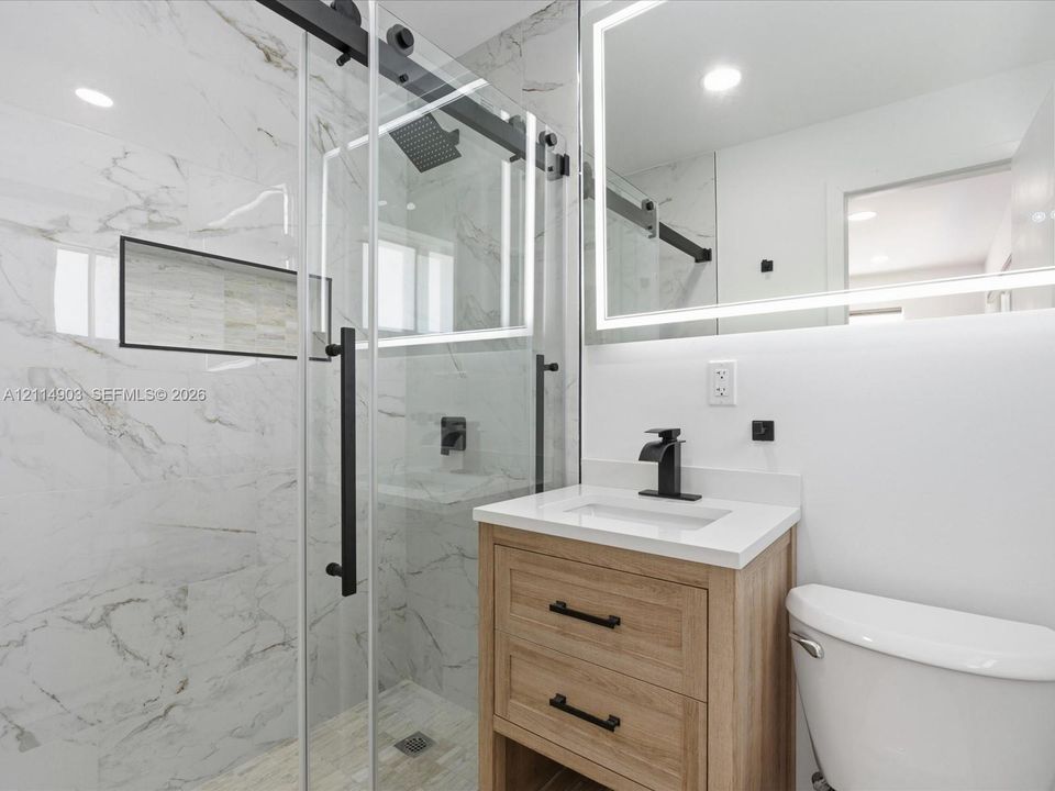 Bathroom featuring a sliding glass shower door with matte black hardware, stone-finish wall tile with integrated recessed shelving, a light wood-finish vanity, a quartz countertop, and an LED-backlit mirror