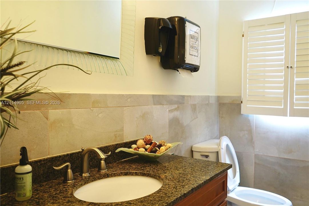 Stone countertop with an undermount sink, natural stone wall tiling, custom wood-finish cabinetry, and white louvered shutters