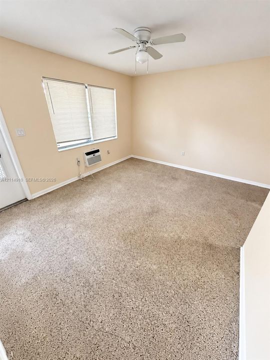 Neutral-toned interior featuring wall-to-wall carpeting, a ceiling fan with integrated lighting, dual-pane windows with horizontal blinds, and a wall-mounted air conditioning unit