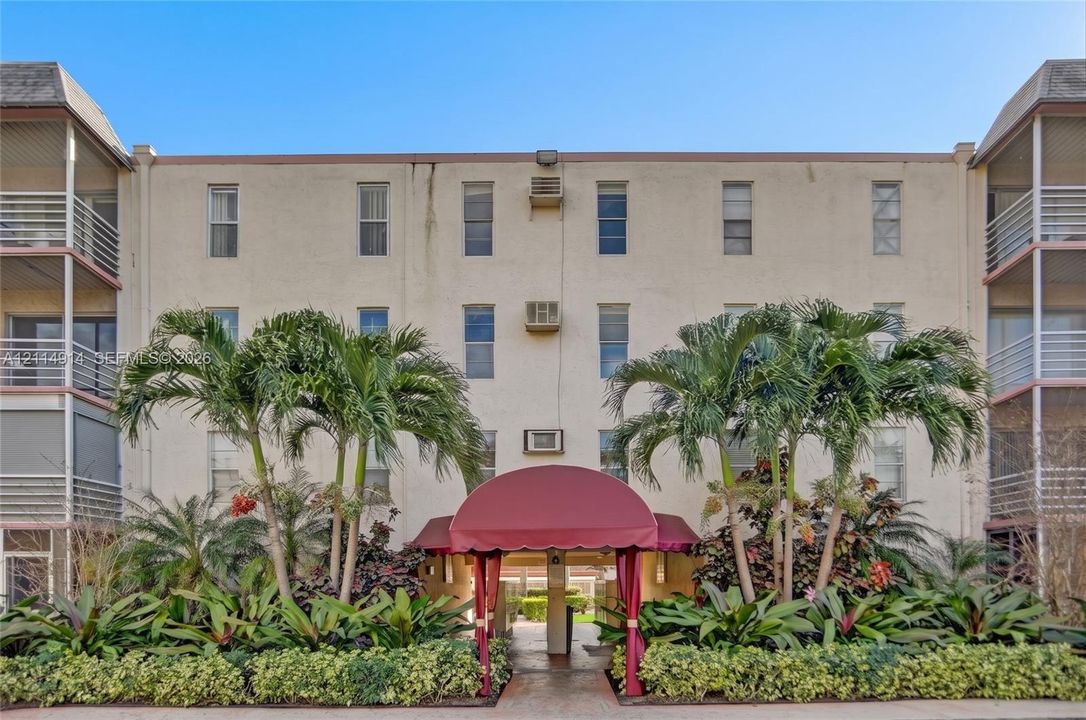 Multi-story stucco exterior featuring stacked balconies with metal railings, a covered arched entryway with fabric awning, and lush tropical landscaping