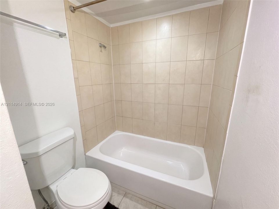 Full-size bathtub with neutral square tile surround, white porcelain toilet, and wall-mounted metal hardware