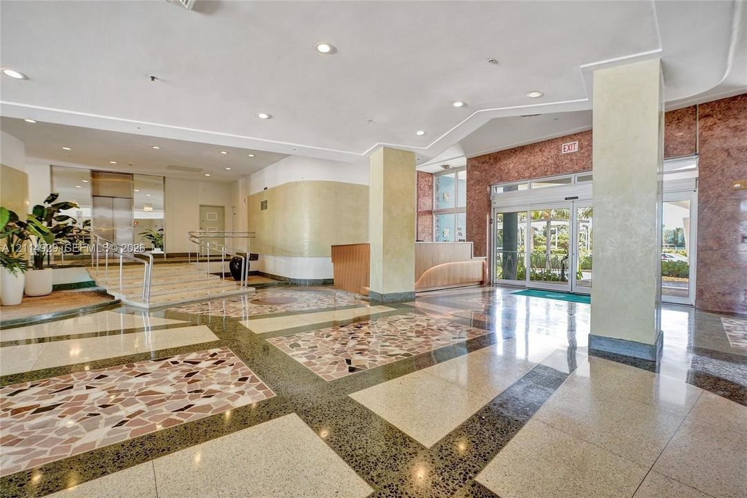 Grand lobby featuring multi-textured stone flooring, high ceilings with recessed lighting, structural support columns, a built-in reception desk, and glass-paned sliding entry doors