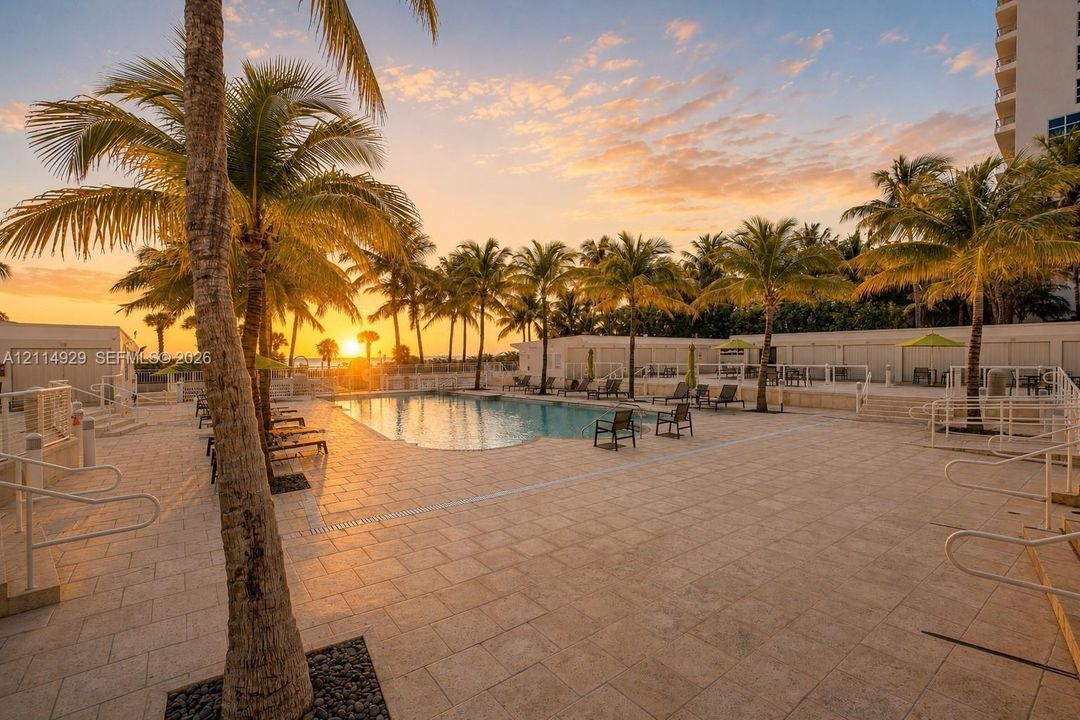 Swimming pool area featuring expansive stone tile decking, mature palm trees, perimeter metal fencing, and integrated pool entry stairs, set against a sunset backdrop