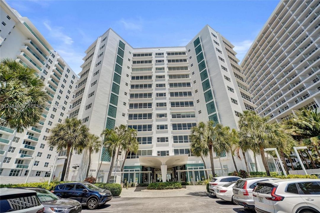 High-rise residential exterior featuring a white concrete facade, multi-story glass-enclosed balconies, and a distinctive structural porte-cochère entry