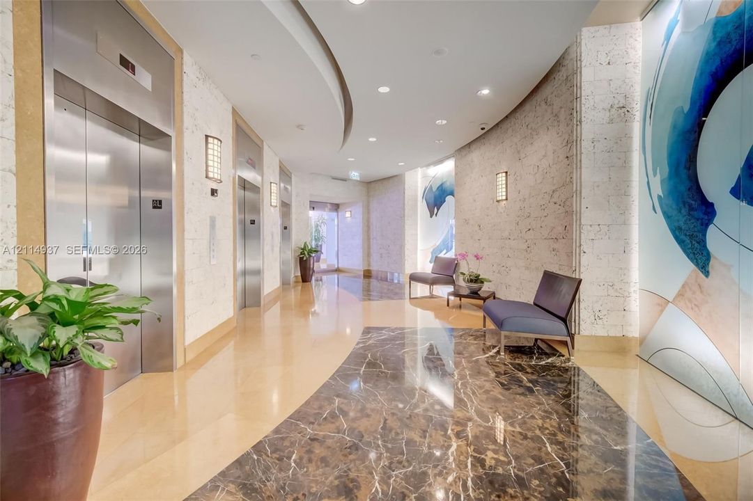 Curved corridor featuring stainless steel elevator doors, polished stone flooring, textured stone wall cladding, and recessed ceiling lighting