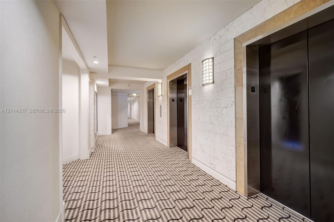 Hallway featuring metallic elevator doors, stone-textured wall cladding, recessed lighting, and patterned carpeting