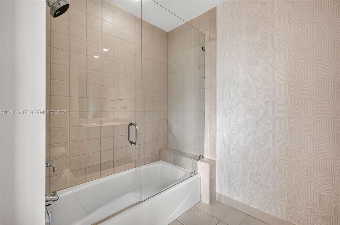 Bathtub with a frameless glass panel, square-tile surround, metallic shower head, and textured accent wall
