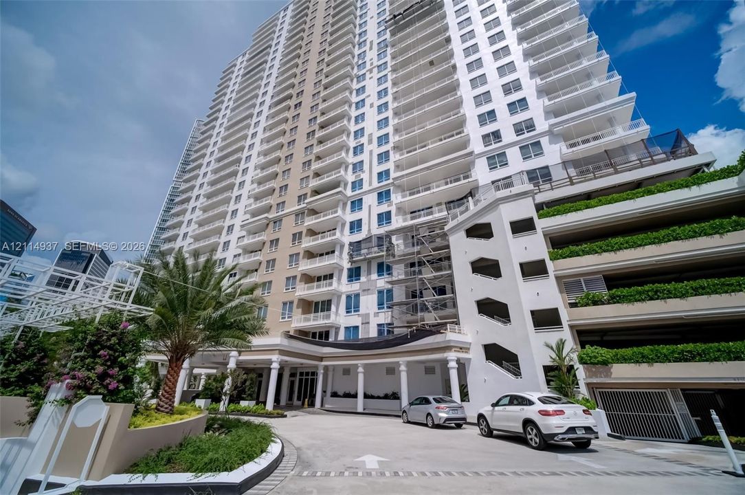 High-rise residential exterior featuring a covered porte-cochère with column supports, tiered balconies, and integrated multi-level parking