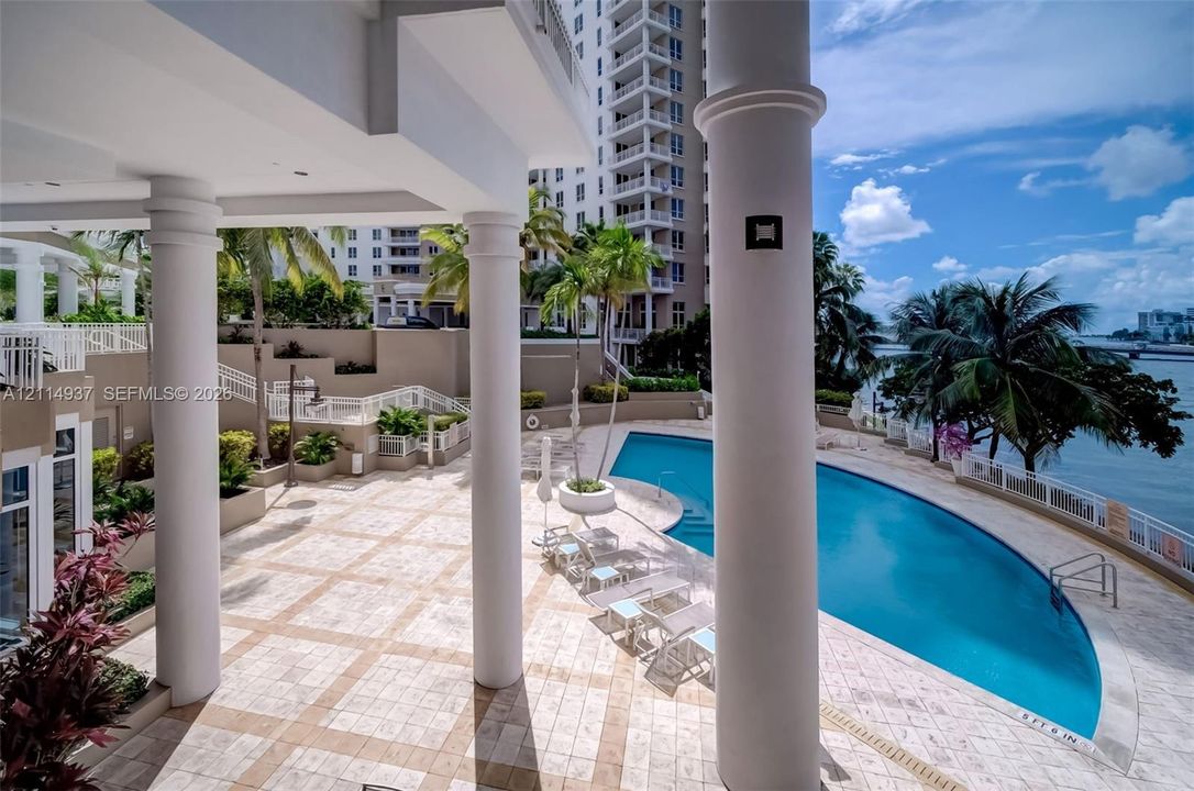 Covered patio featuring large structural columns, expansive stone-tiled decking, a curved swimming pool, and direct water frontage