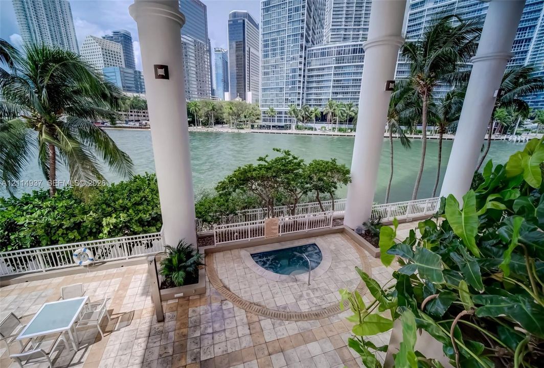 Covered terrace featuring full-height architectural columns, tiled stone flooring, a built-in spa, and protective white metal fencing