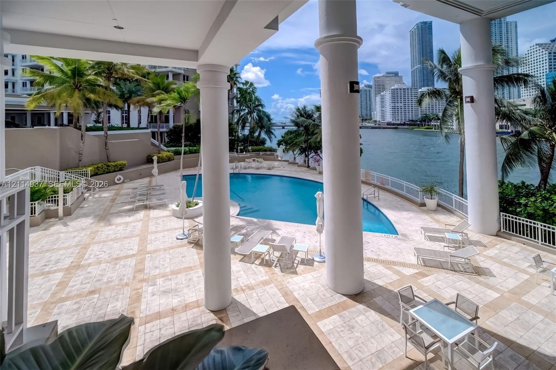 Covered terrace featuring structural columns, stone-tiled patio, and an in-ground pool situated along the waterfront