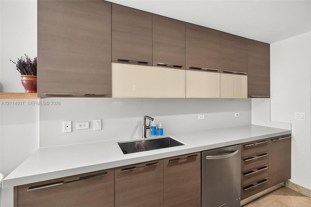 Contemporary cabinetry featuring wood-finish and high-gloss cream flat-panel doors, solid-surface countertops, integrated stainless steel sink with a pull-down faucet, and built-in stainless steel dishwasher