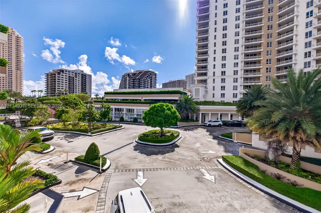 Paved arrival court featuring a landscaped central island, manicured hedging, established palm trees, and structural concrete high-rise exterior