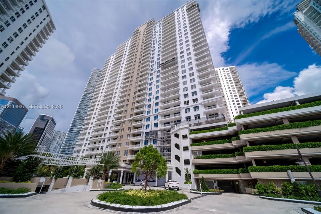 High-rise residential tower featuring tiered concrete balconies, covered multi-level parking structure, integrated landscape planters, and a ground-level porte-cochere