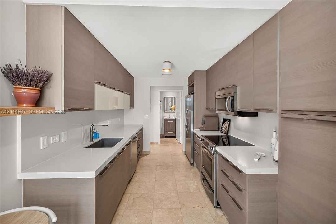 Galley kitchen featuring flat-panel wood-finish cabinetry, white solid-surface countertops, stone tile flooring, and integrated stainless steel appliances