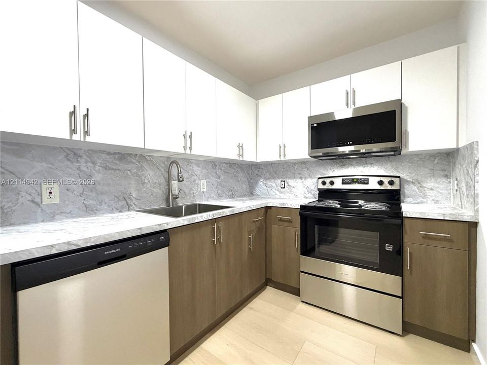 Kitchen featuring two-tone flat-panel cabinetry, light-toned stone-patterned countertops and backsplash, stainless steel appliances, and wood-finish flooring