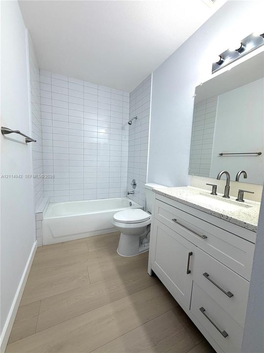Bathroom featuring wood-finish flooring, white cabinetry with stone-composite countertops, and a white tiled tub-shower surround