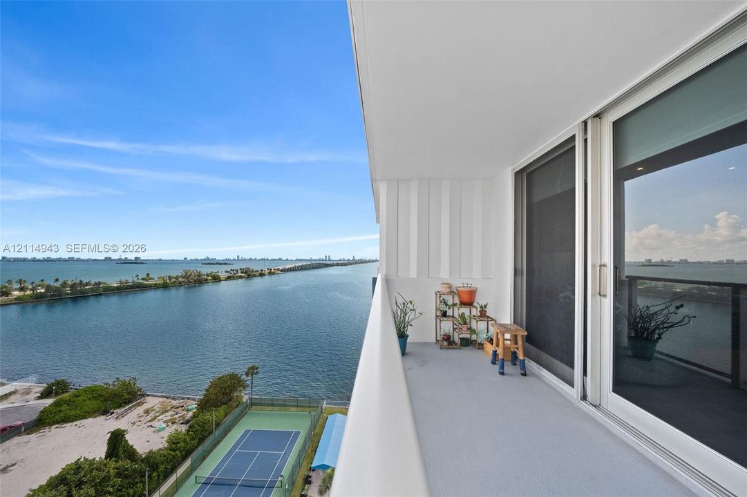 Private balcony with floor-to-ceiling sliding glass doors and concrete flooring, featuring an expansive water panorama and a hard-surface tennis court