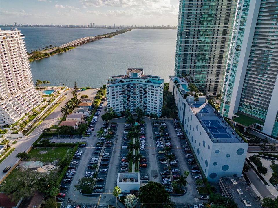 Aerial perspective of waterfront mid-rise building featuring structured parking, rooftop tennis court, and proximity to the bay and causeway