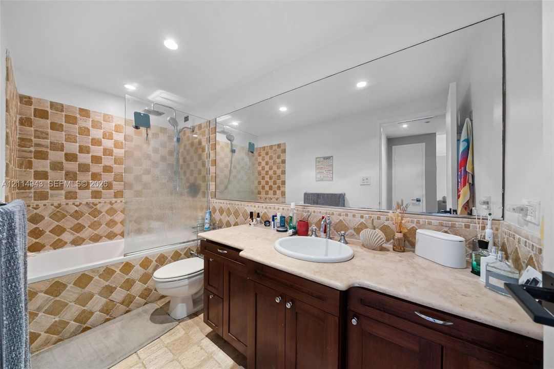 Bathroom featuring dark wood cabinetry, a stone-finish countertop with an integrated oval sink, a wall-to-wall mirror, and decorative tile accents surrounding the bathtub