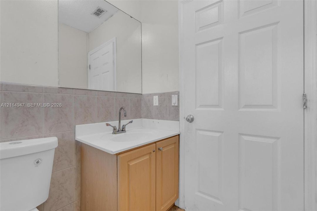 Vanity featuring a light wood finish cabinet, white solid-surface countertop, integrated sink, and dual-handle faucet, complemented by a large wall-mounted mirror and neutral-toned tile wainscoting