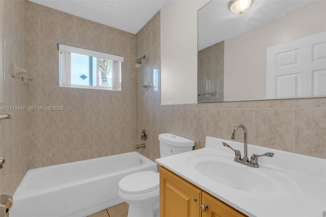 Bathroom featuring a wood-finish vanity with a white countertop, an integrated sink with a dual-handle faucet, a full-width wall mirror, and neutral-toned wall tiling surrounding a standard tub-shower combination