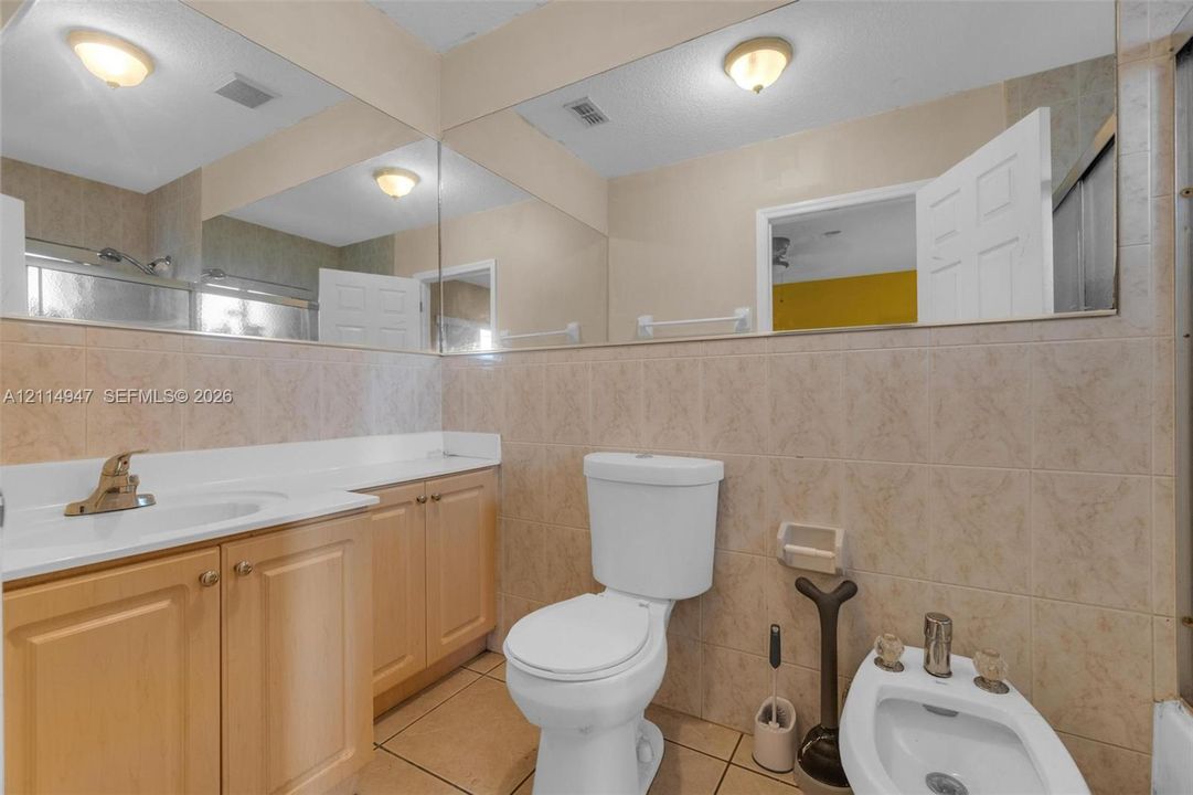 Bathroom featuring a wood-finish vanity with a white countertop, full-width wall mirror, ceramic wall tiling, and a bidet