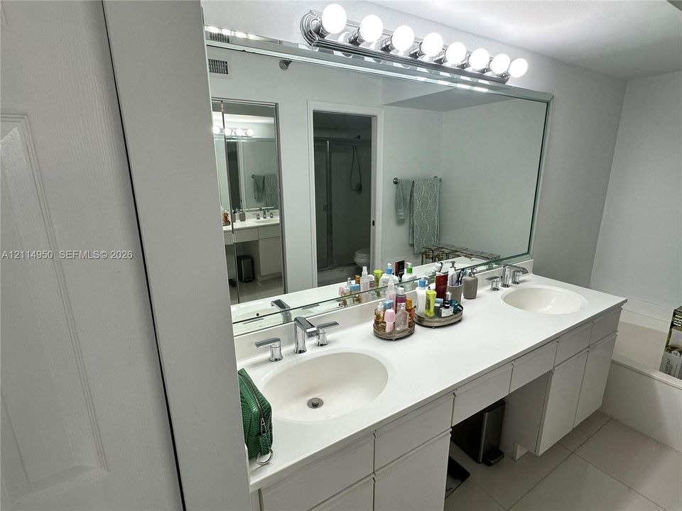 Dual-sink vanity featuring a solid surface countertop, integrated basins, chrome hardware, and a large-format vanity mirror with an attached eight-bulb light fixture
