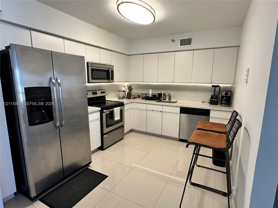 Kitchen featuring white flat-panel cabinetry, stainless steel appliances, solid-surface countertops, integrated under-cabinet lighting, and large-format tile flooring