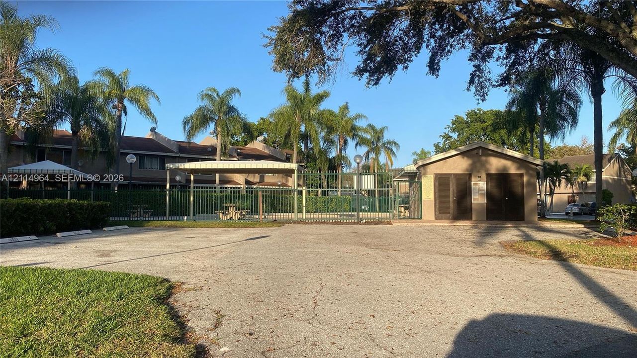 Community pool area featuring perimeter fencing, a covered canopy structure, a stand-alone utility building, and mature palm trees