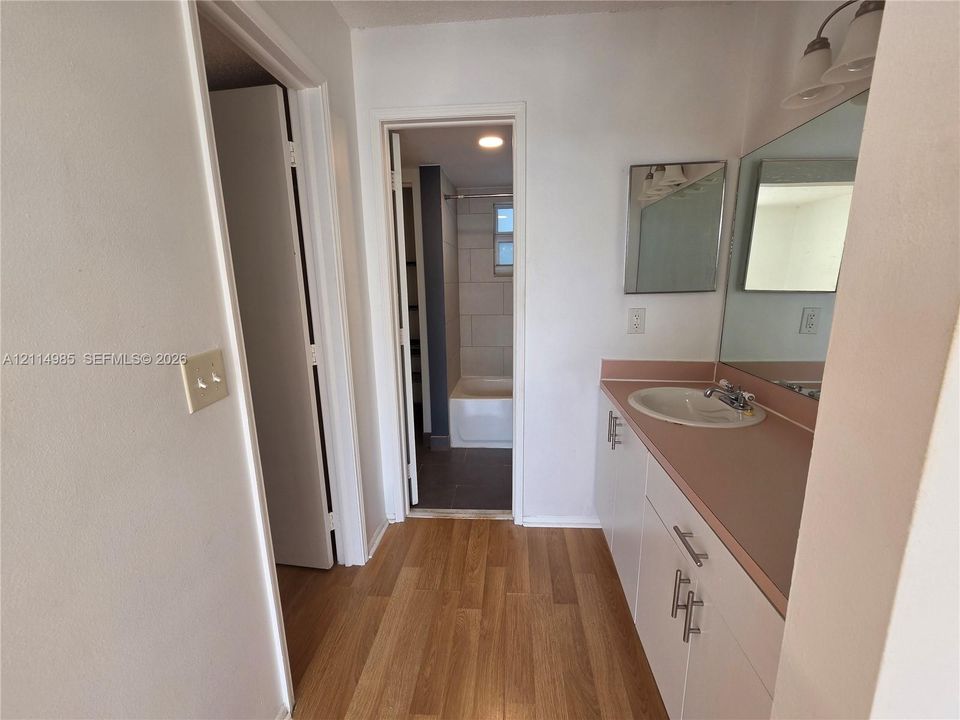 Bathroom featuring wood-finish flooring, a white vanity with a solid-surface countertop and integrated sink, a full-width wall mirror, and a tiled shower enclosure