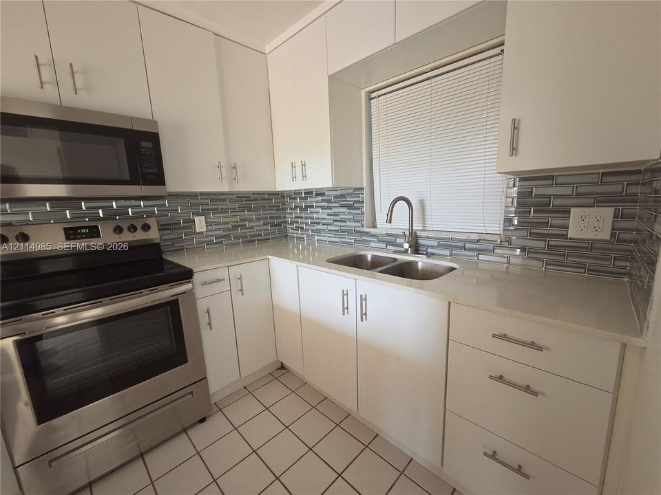 Kitchen featuring white flat-panel cabinetry, stainless steel appliances, light-toned stone countertops, a multi-tonal glass tile backsplash, and dual-basin sink with a gooseneck faucet
