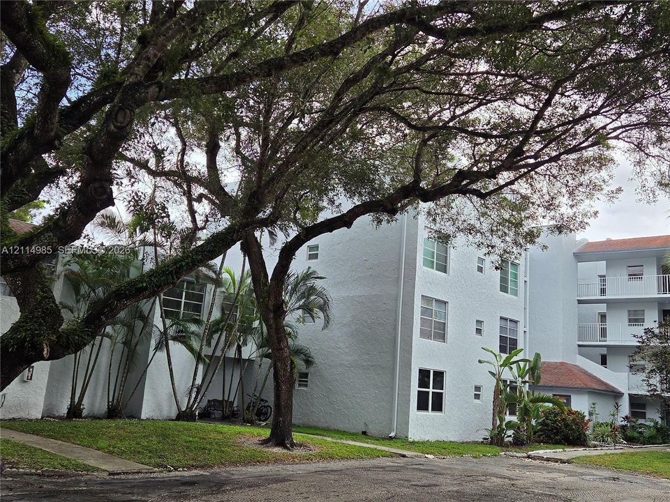 Multi-story building featuring textured stucco exterior, grid-pattern windows, multi-level exterior balconies with white railings, and mature trees